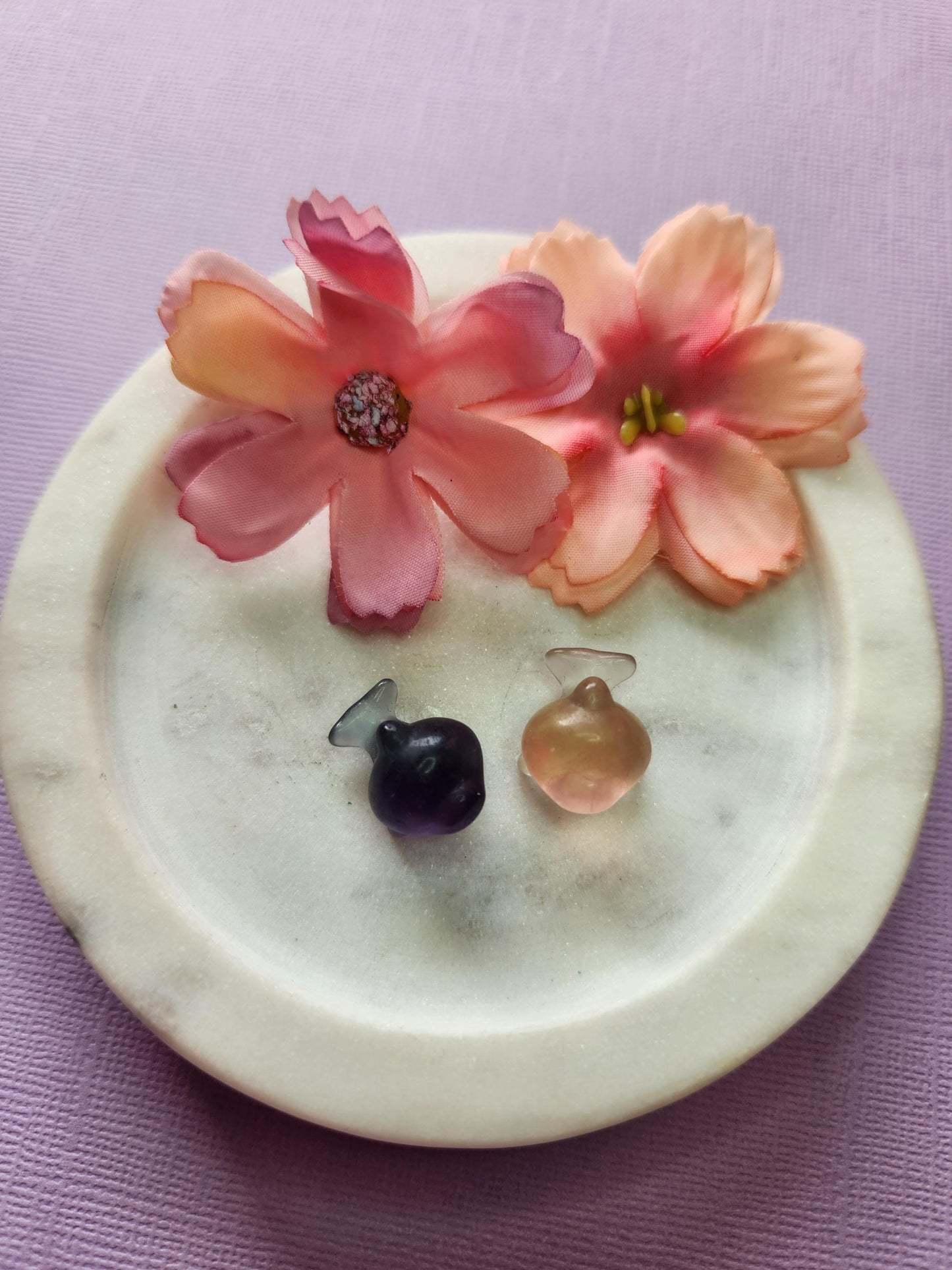 Close-up photo of two fluorite mini whale carvings displayed in a white dish with two soft flower props on a lavender backdrop. The whales show natural bands of purple, green and blue fluorite with a glossy polished finish. Ideal for fluorite carvings, mini crystal animals, whale crystal decor and collectible gemstone figures