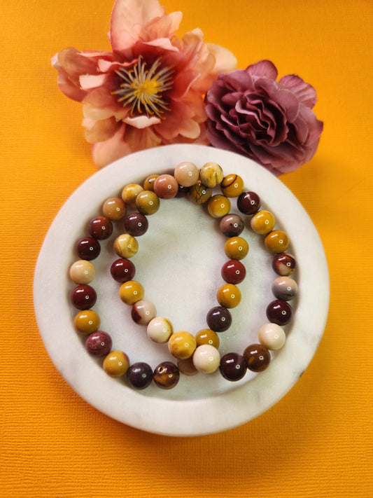 Two Mookaite 8mm beaded bracelets in a white dish with two flower props on a yellow background, natural Mookaite gemstone beads, handmade crystal jewellery, grounding and energizing stone bracelet product photo for Etsy and Shopify listings.
