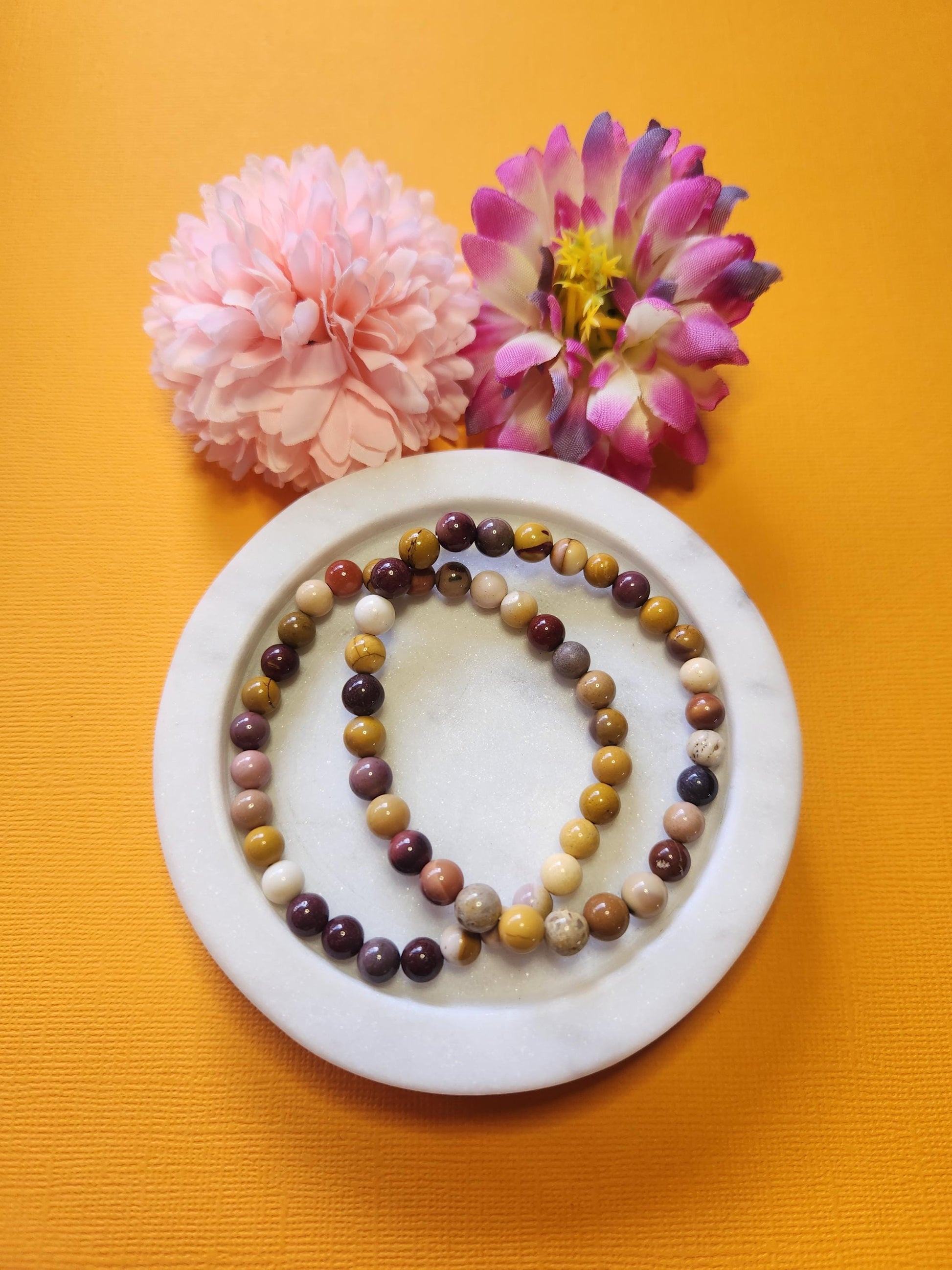 Two Mookaite 6mm beaded crystal bracelets displayed on a white dish with two flower props against a vibrant yellow background. The natural red, mustard, and cream tones of the Mookaite jasper beads are shown in detail, highlighting their earthy Australian origin. Perfect for grounding, confidence, and emotional balance, this handmade Mookaite bracelet adds vibrant crystal energy to any boho or spiritual jewelry collection.