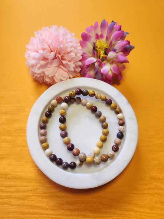 Two Mookaite 6mm beaded crystal bracelets displayed on a white dish with two flower props against a vibrant yellow background. The natural red, mustard, and cream tones of the Mookaite jasper beads are shown in detail, highlighting their earthy Australian origin. Perfect for grounding, confidence, and emotional balance, this handmade Mookaite bracelet adds vibrant crystal energy to any boho or spiritual jewelry collection.