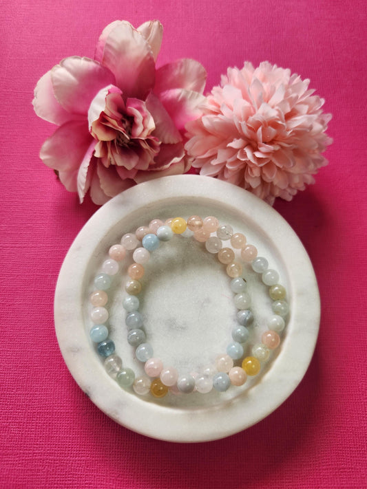 Two Morganite 6mm beaded bracelets displayed on a white dish with two flower props against a bright pink background. The soft pastel gemstones showcase gentle pink and peach hues, radiating loving, calming, and heart-healing energy. Perfect for emotional balance, self-love rituals, and crystal jewelry collections.