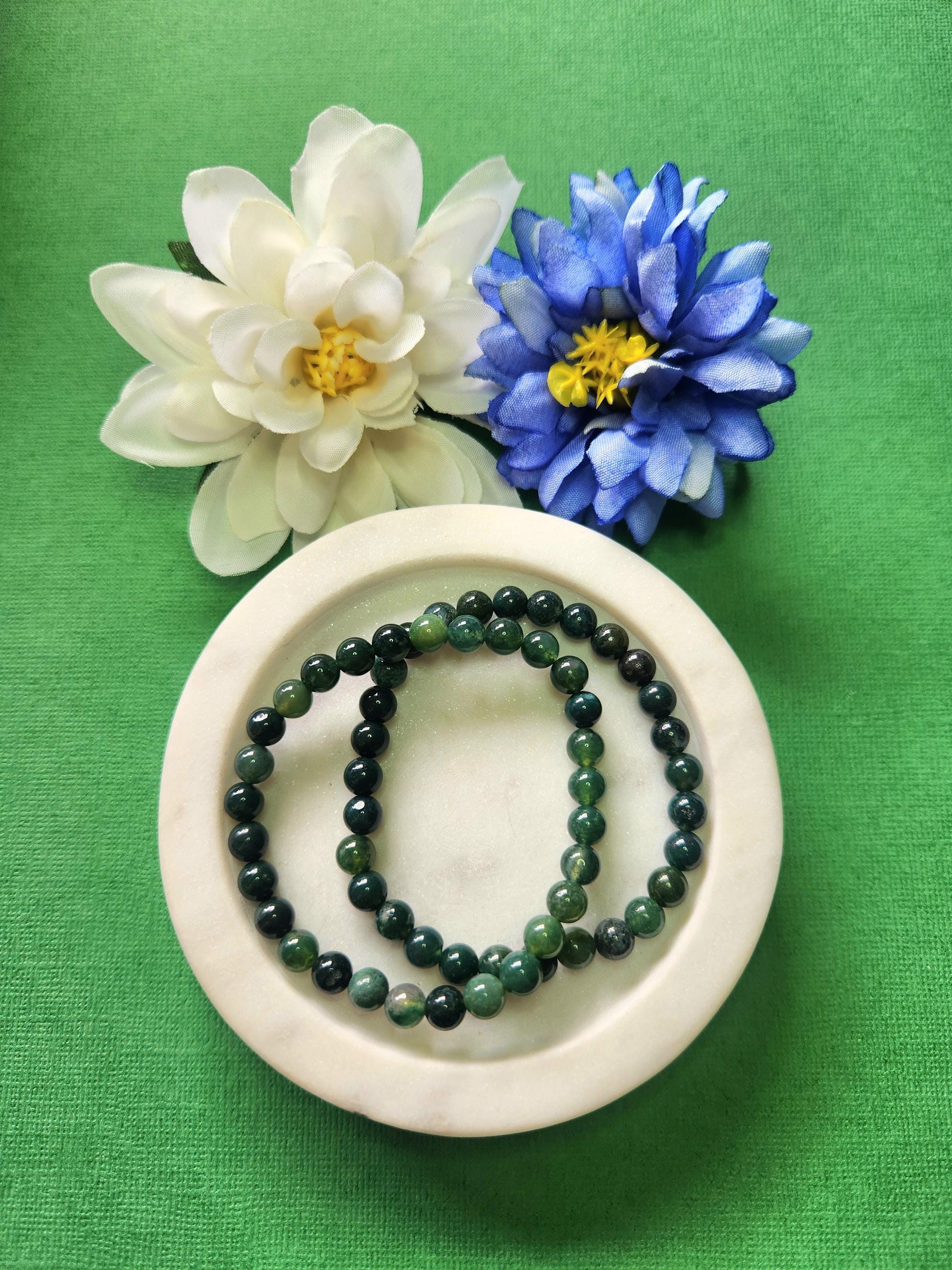 Two Moss Agate 6mm beaded bracelets displayed on a white dish with two flower props against a green background. The natural green and white marbled gemstones radiate grounding, growth, and abundance energy. Perfect for nature lovers, spiritual grounding, and crystal healing jewelry collections.