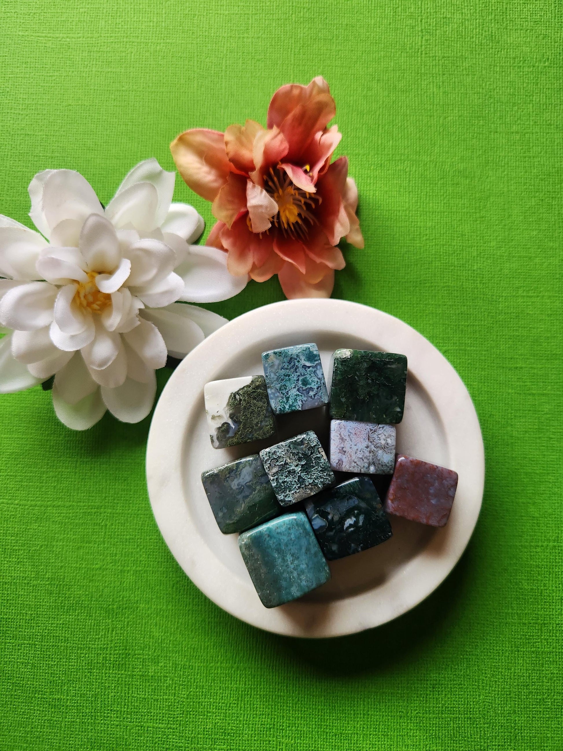 Moss Agate cubes displayed on a green background, showcasing their natural green and white patterns that resemble forest moss. These polished gemstone cubes are known for grounding energy, connection to nature, and heart chakra healing. Ideal for meditation, crystal grids, and spiritual decor. The green backdrop enhances the earthy tones and organic beauty of the stones, making them perfect for collectors and energy workers.