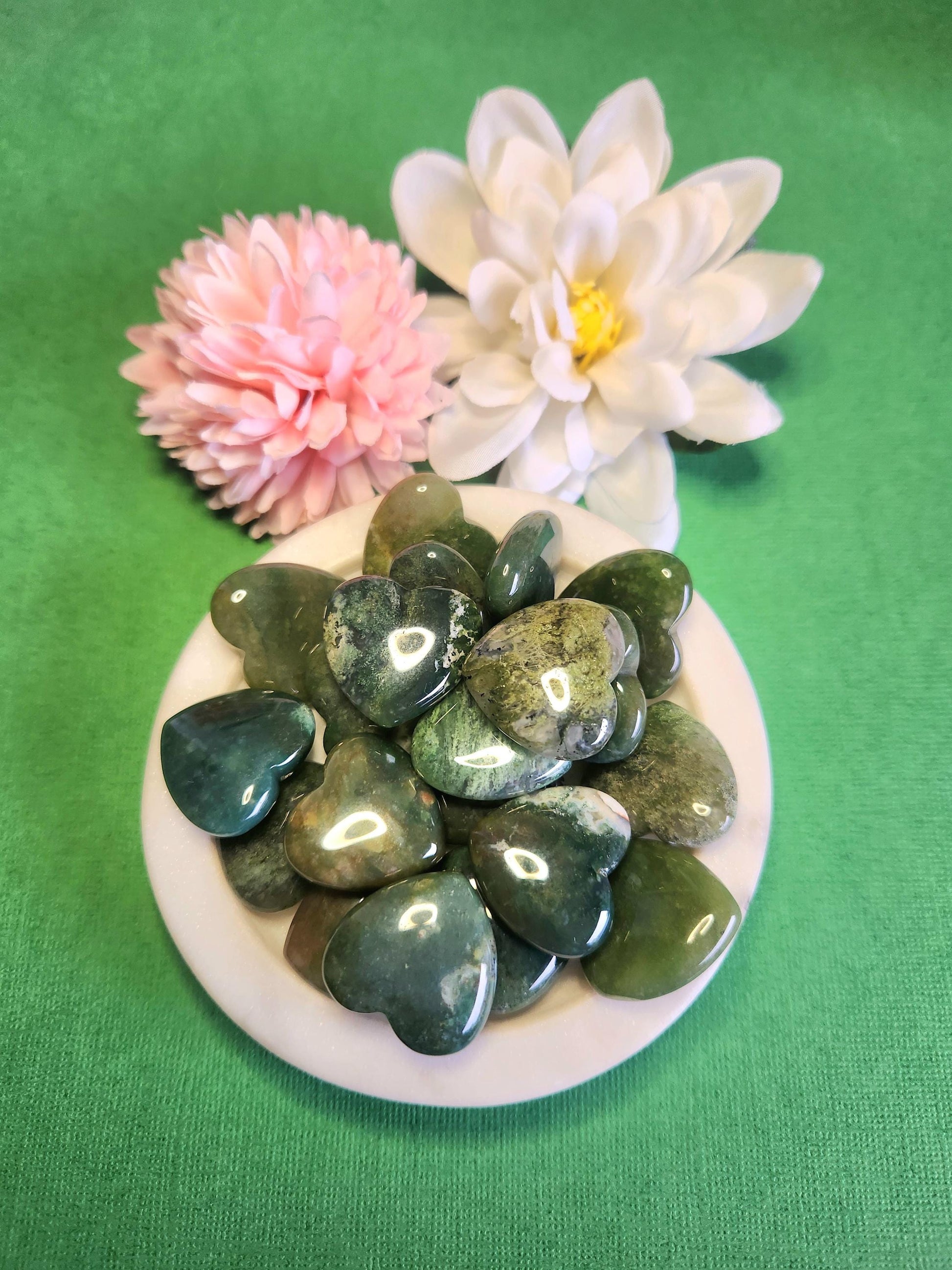 Moss agate crystal heart displayed in a white dish on a green background with soft flower props featuring polished heart stone for growth balance grounding healing crystal decor and gift
