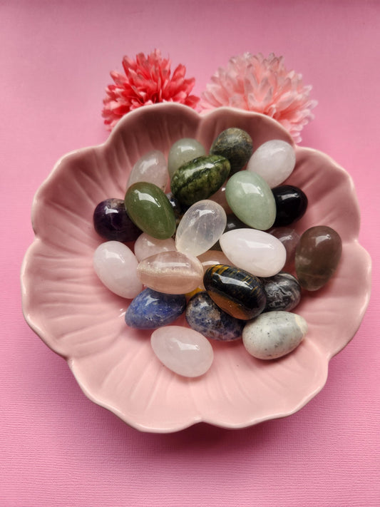 Mystery crystal eggs featuring mixed gemstone crystal egg carvings in assorted colors. Polished crystal eggs styled in a pink flower bowl with decorative flower props. Pink background product photo for mystery crystals, gemstone decor, and crystal gifts.