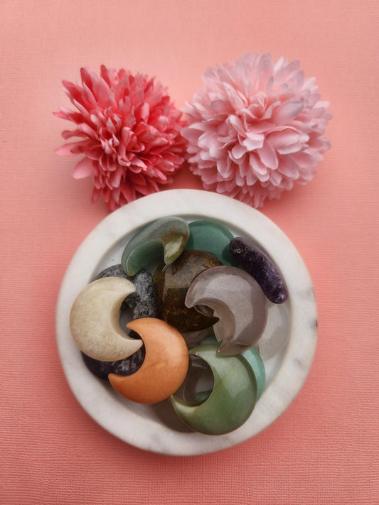 Mystery crystal moon carvings displayed in a white ceramic dish with two floral props on a warm orange background, featuring assorted natural healing crystals and gemstones, crescent moon crystal carvings, metaphysical crystals for intuition manifestation and spiritual energy, small crystal moons perfect for gifting altar decor and crystal collections