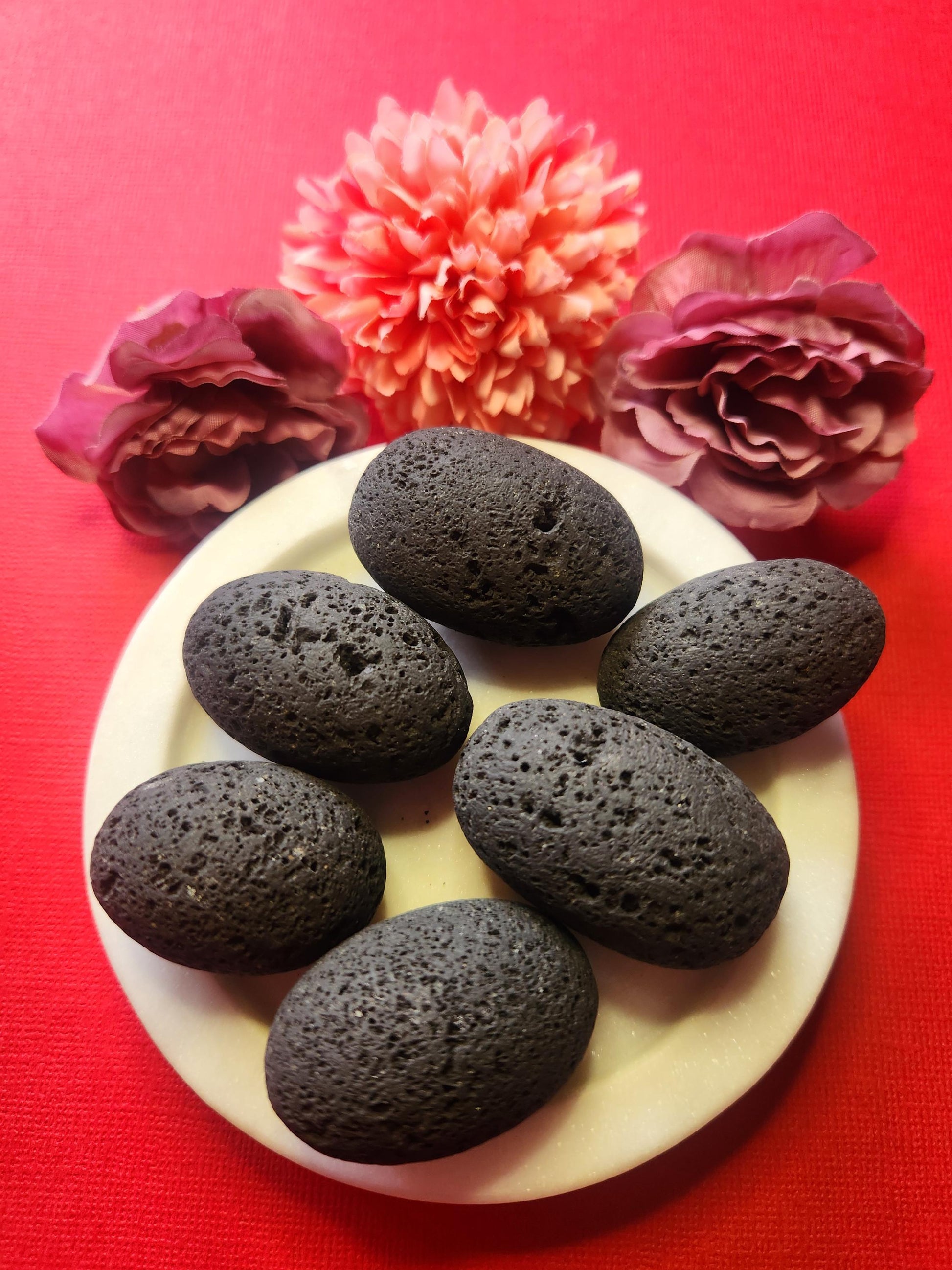 Lava stone tumbles in a white dish with two flower props on a red background, natural volcanic healing stones for grounding, protection, and aromatherapy use, high-quality crystal tumbles product photo for shop listing