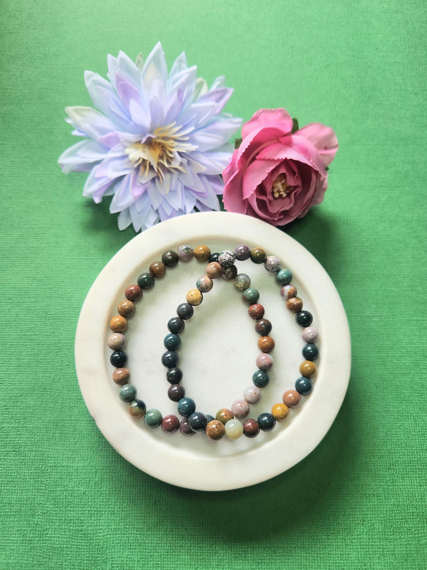 Two Ocean Jasper 6mm beaded bracelets displayed on a white dish with two flower props against a green background. The natural green, cream, and earthy patterned gemstones radiate calming, joyful, and balancing energy. Perfect for emotional healing, meditation, and crystal jewelry collections.
