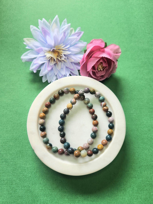 Two Ocean Jasper 6mm beaded bracelets displayed on a white dish with two flower props against a green background. The natural green, cream, and earthy patterned gemstones radiate calming, joyful, and balancing energy. Perfect for emotional healing, meditation, and crystal jewelry collections.