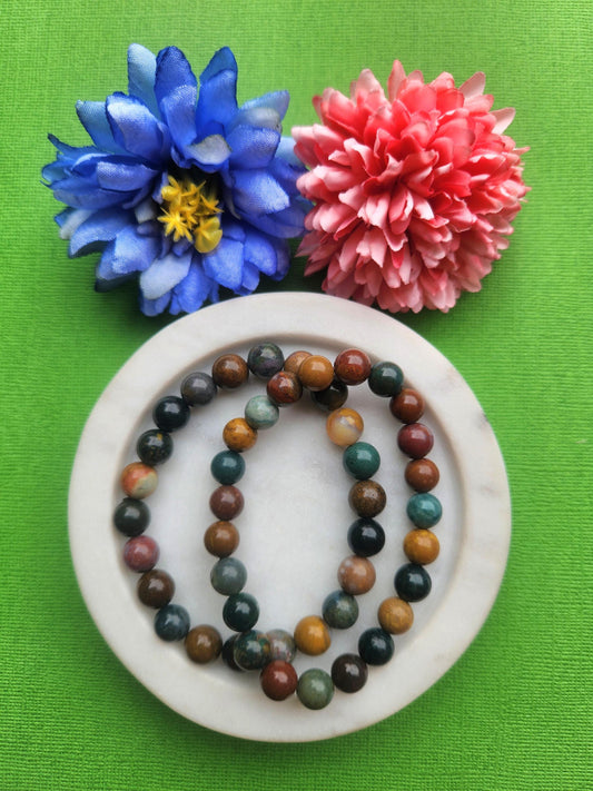 Two ocean jasper 8mm beaded bracelets displayed in a white dish on a green background with soft flower props featuring polished crystal beads for calm joy emotional balance healing energy bracelet and gift