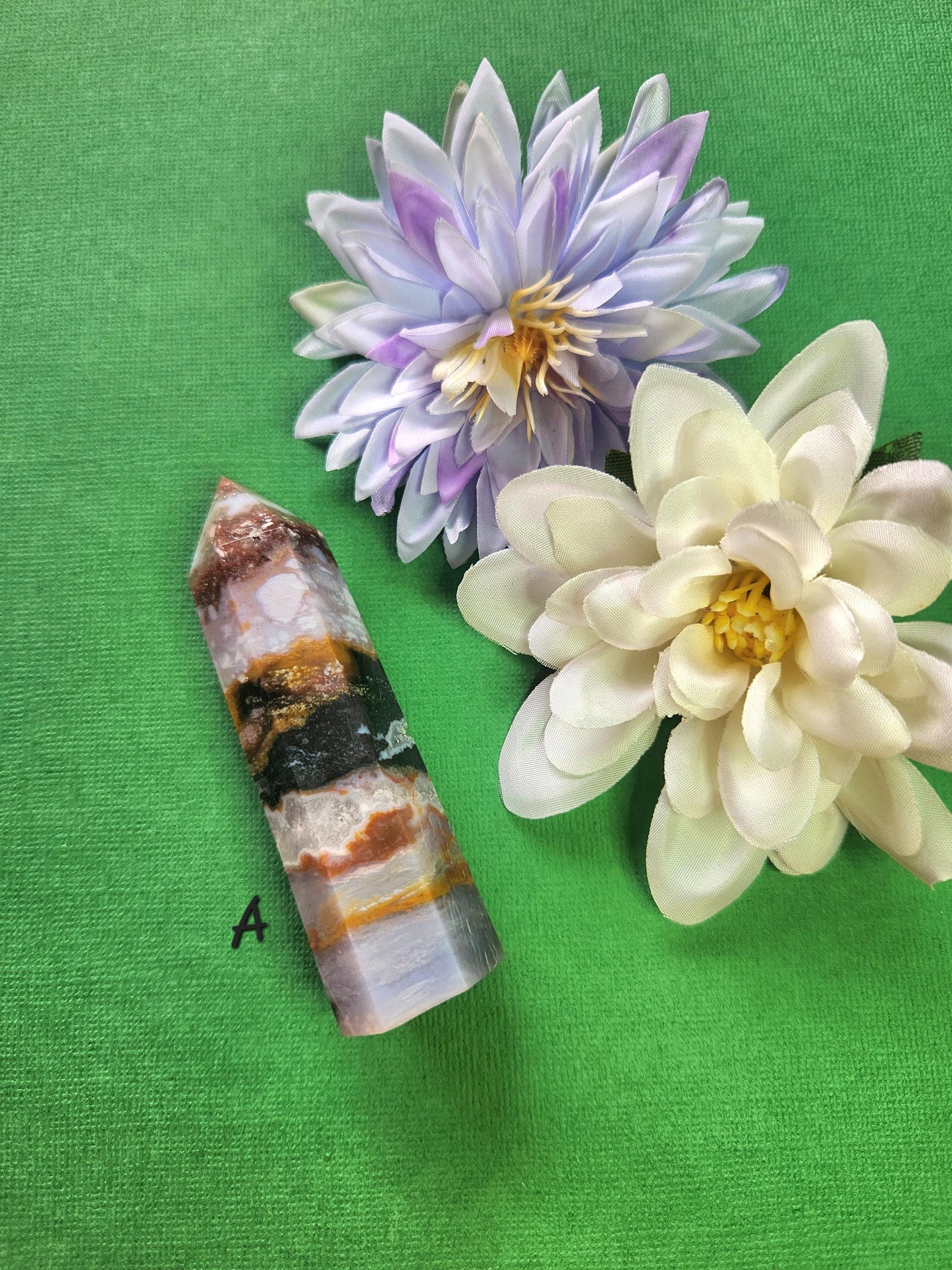 Polished Ocean Jasper tower A with green, cream, and brown orbicular patterns, displayed with two flower props on a green background, natural gemstone point for meditation, energy balancing, spiritual healing, and crystal collection décor