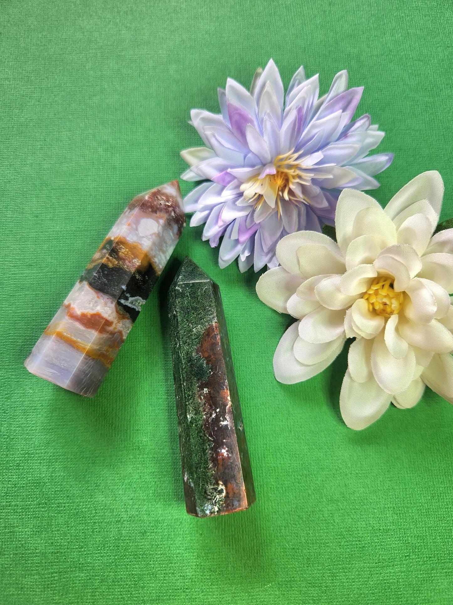 Two polished Ocean Jasper crystal towers with natural green, cream, and brown orbicular patterns, displayed with two flower props on a green background, high-quality gemstone points for emotional healing, grounding, meditation, and metaphysical décor