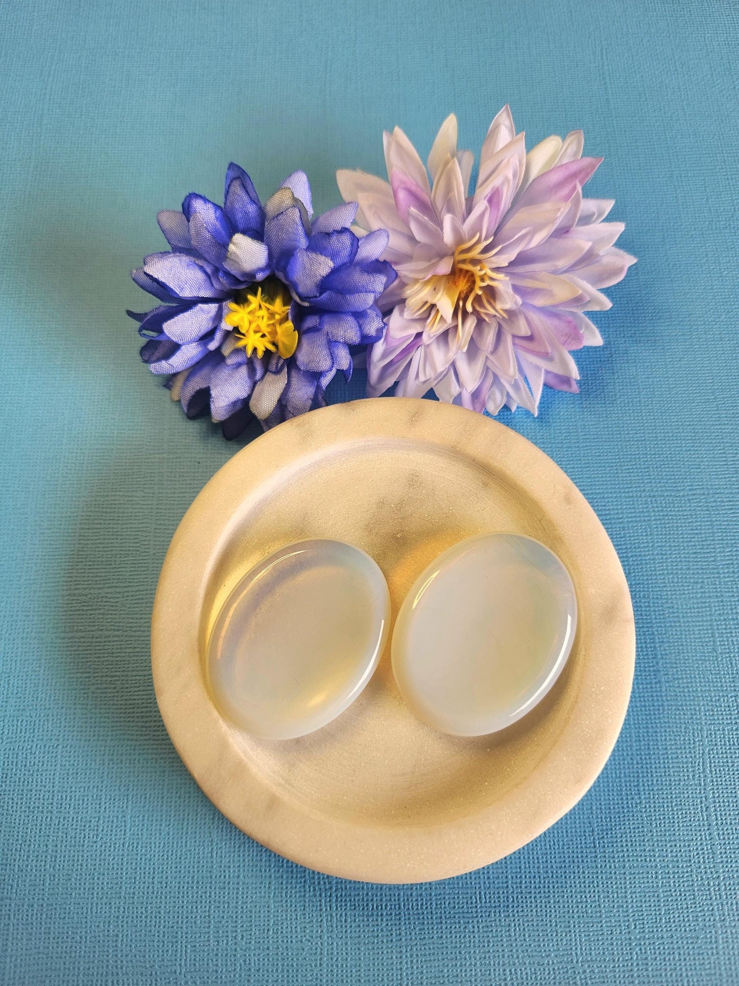 Opalite worry stone displayed in a white dish on a blue background with soft flower props featuring polished opalite stone for calming energy emotional balance clarity stress relief crystal decor and gift