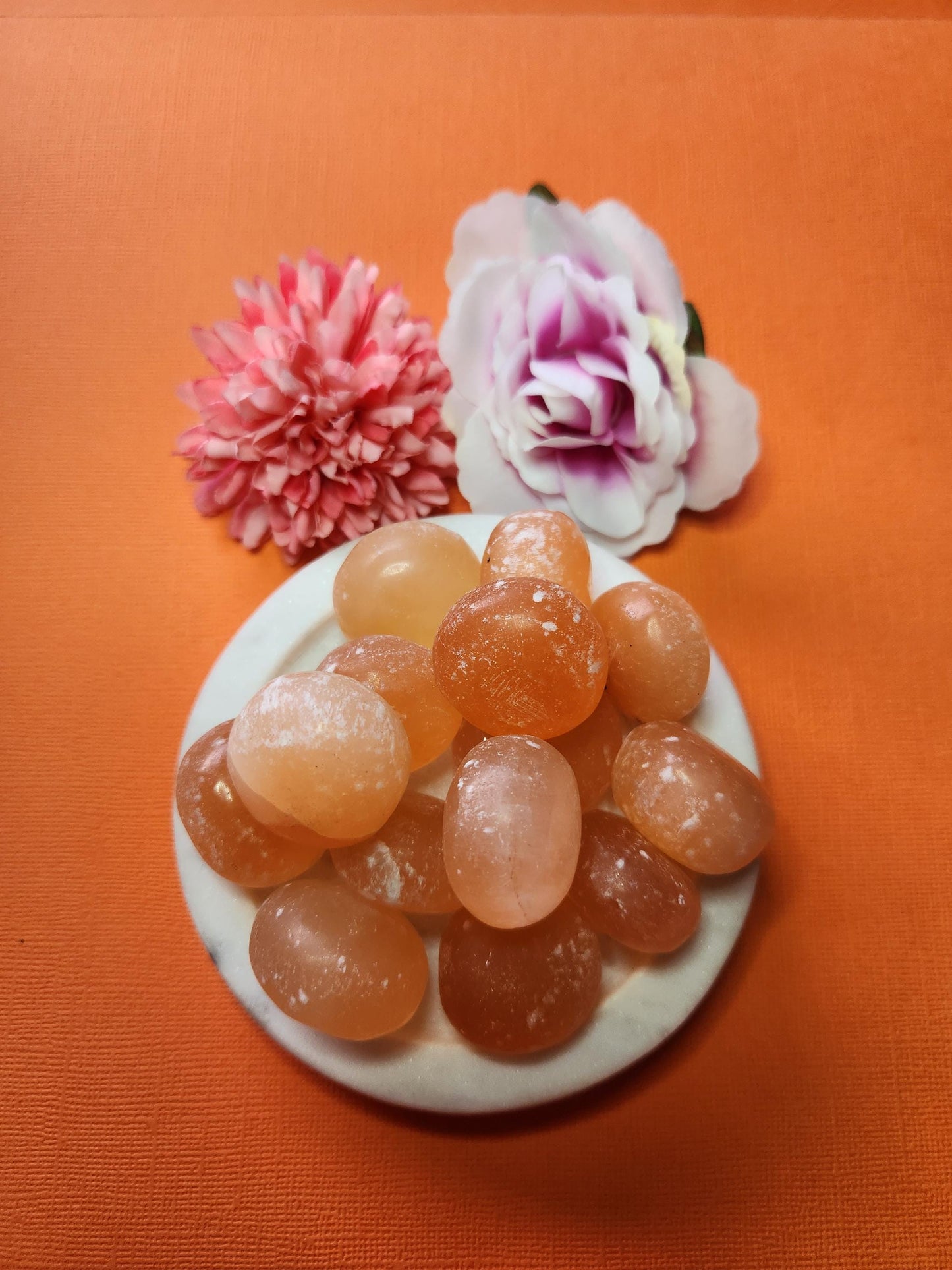 Close-up photo of several peach selenite tumbles displayed in a white dish with two soft flower props on an orange backdrop. The tumbles show soft peach-orange hues with a smooth, polished finish. Ideal for selenite crystals, tumbled gemstones, crystal collections, and decorative crystal accents.