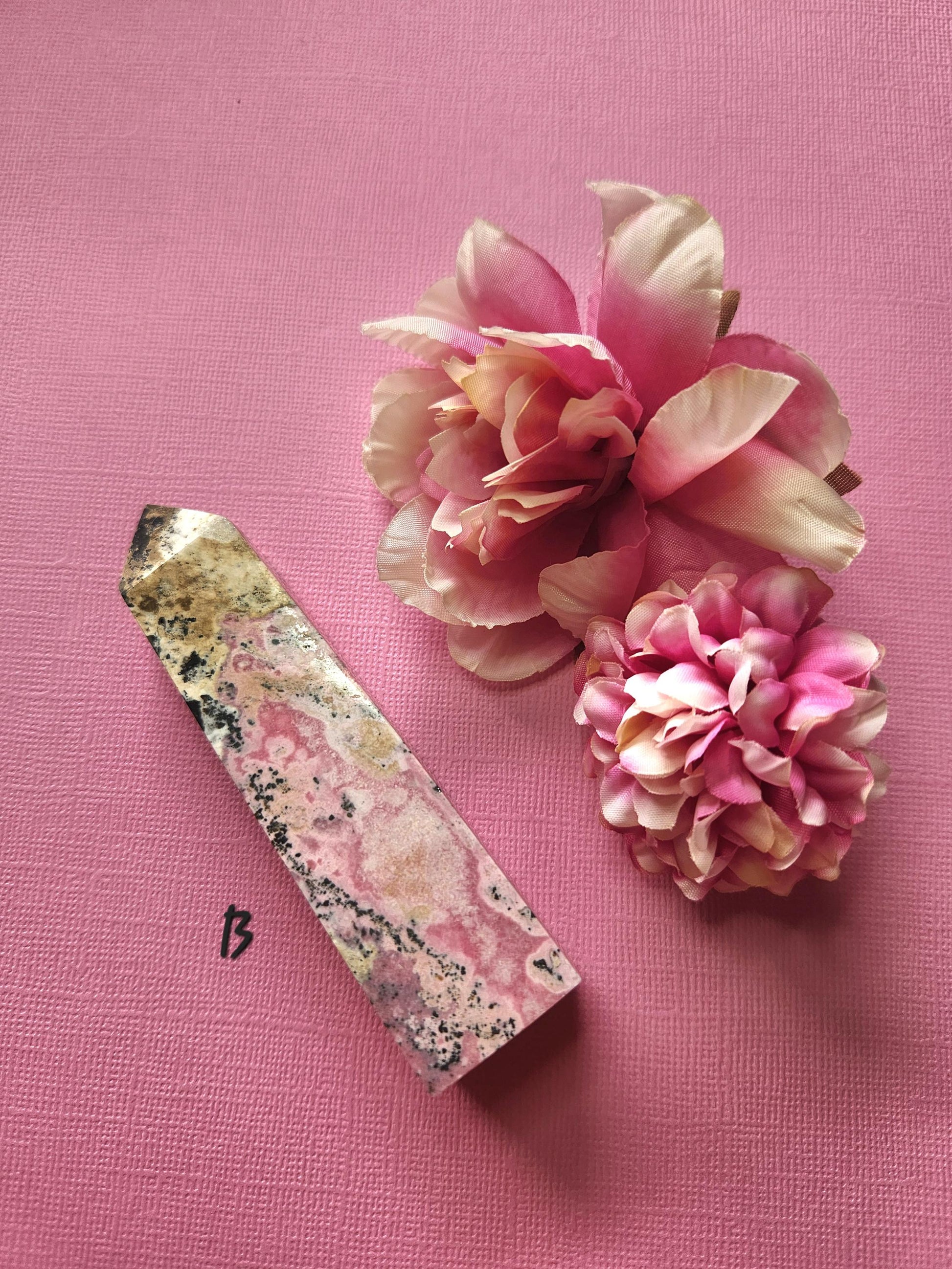 Peruvian Rhodonite tower B captured from different angle highlighting natural pink tones and dark veining, polished crystal point with two flower props on pink background, high-energy self-love rhodonite tower for metaphysical healing