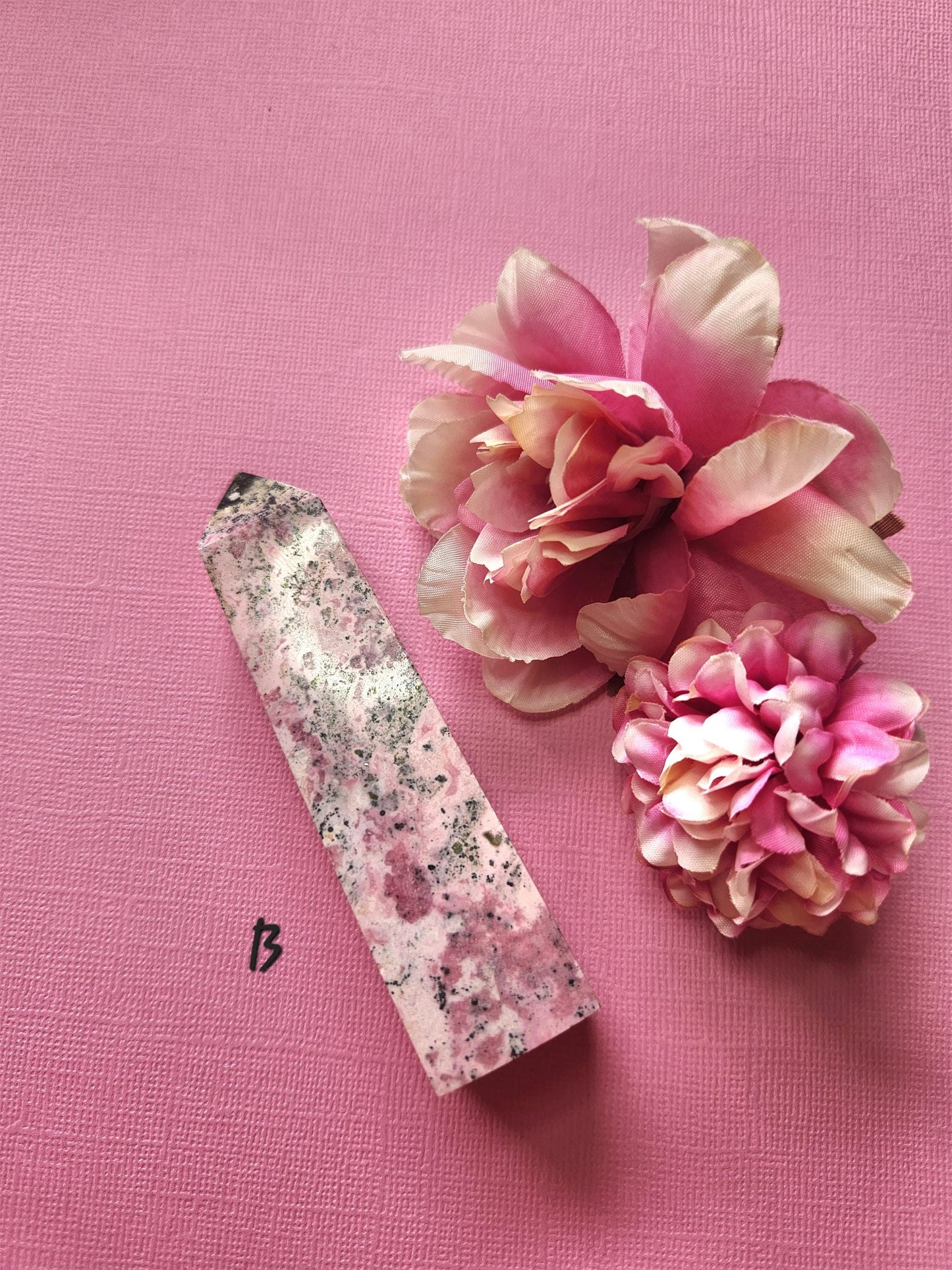 Peruvian Rhodonite tower B with striking pink, charcoal and deep rose patterning, polished crystal point displayed with two flower props on a pink backdrop, premium rhodonite gemstone tower for heart chakra and emotional healing