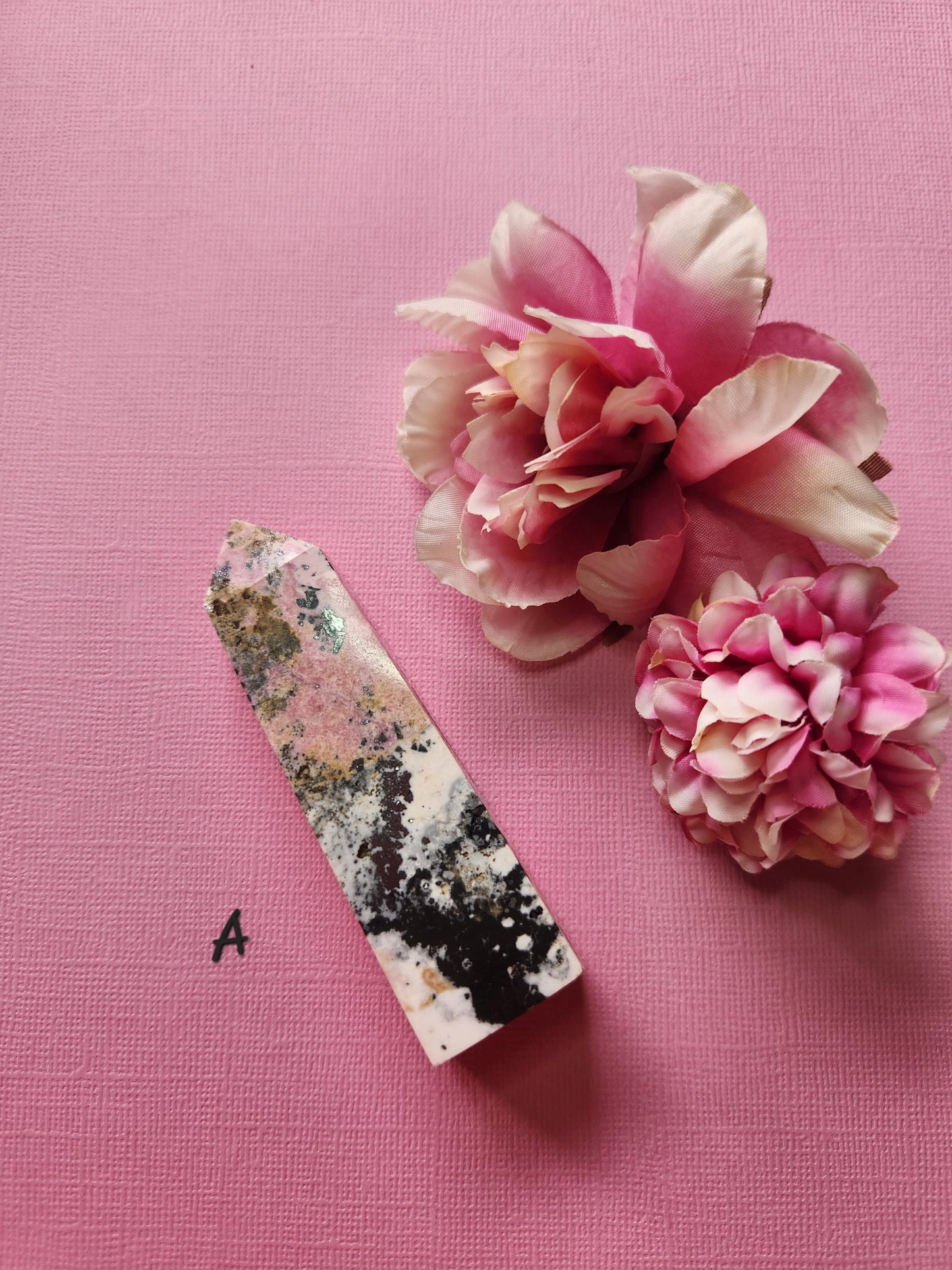 Peruvian Rhodonite tower A with vibrant pink and black veining, polished crystal point displayed with two flower props on a pink backdrop, self-love gemstone tower, healing crystal décor, high-grade rhodonite point