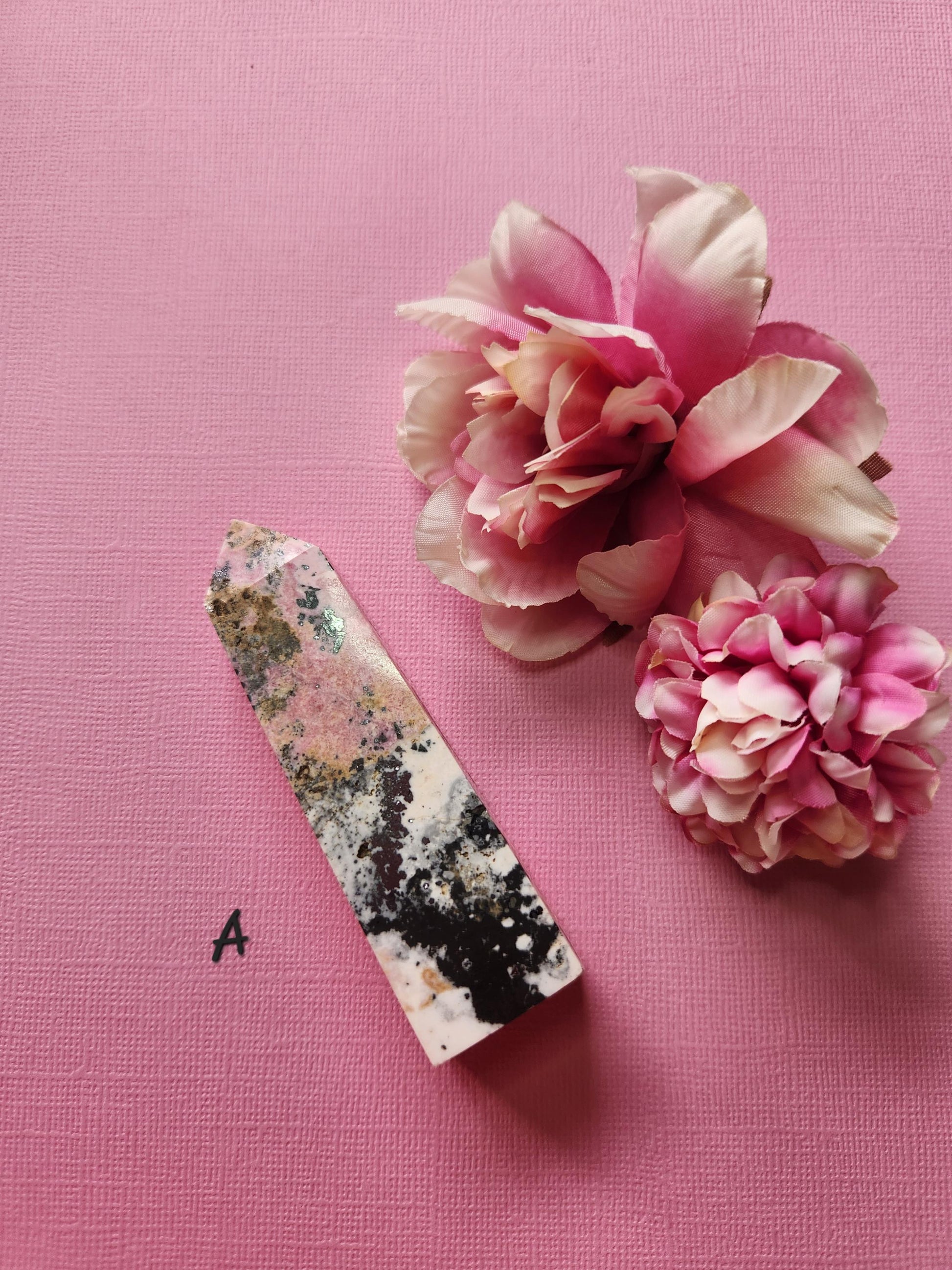 Peruvian Rhodonite tower A with vibrant pink and black veining, polished crystal point displayed with two flower props on a pink backdrop, self-love gemstone tower, healing crystal décor, high-grade rhodonite point