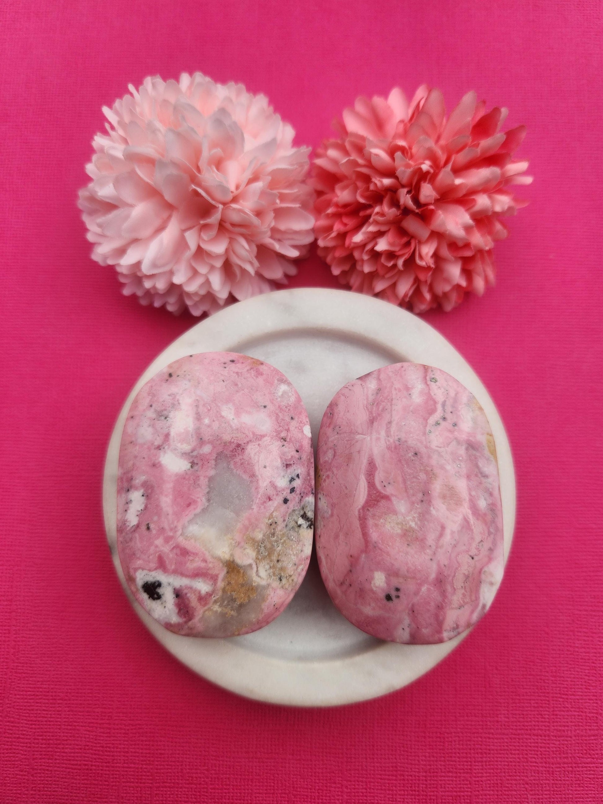 Peruvian rhodonite palmstones displayed in a white ceramic dish with two floral props on a soft pink background, featuring natural rhodonite crystals polished palm stones, healing gemstones for emotional balance self love grounding energy, crystal palmstones perfect for gifting meditation and crystal collections