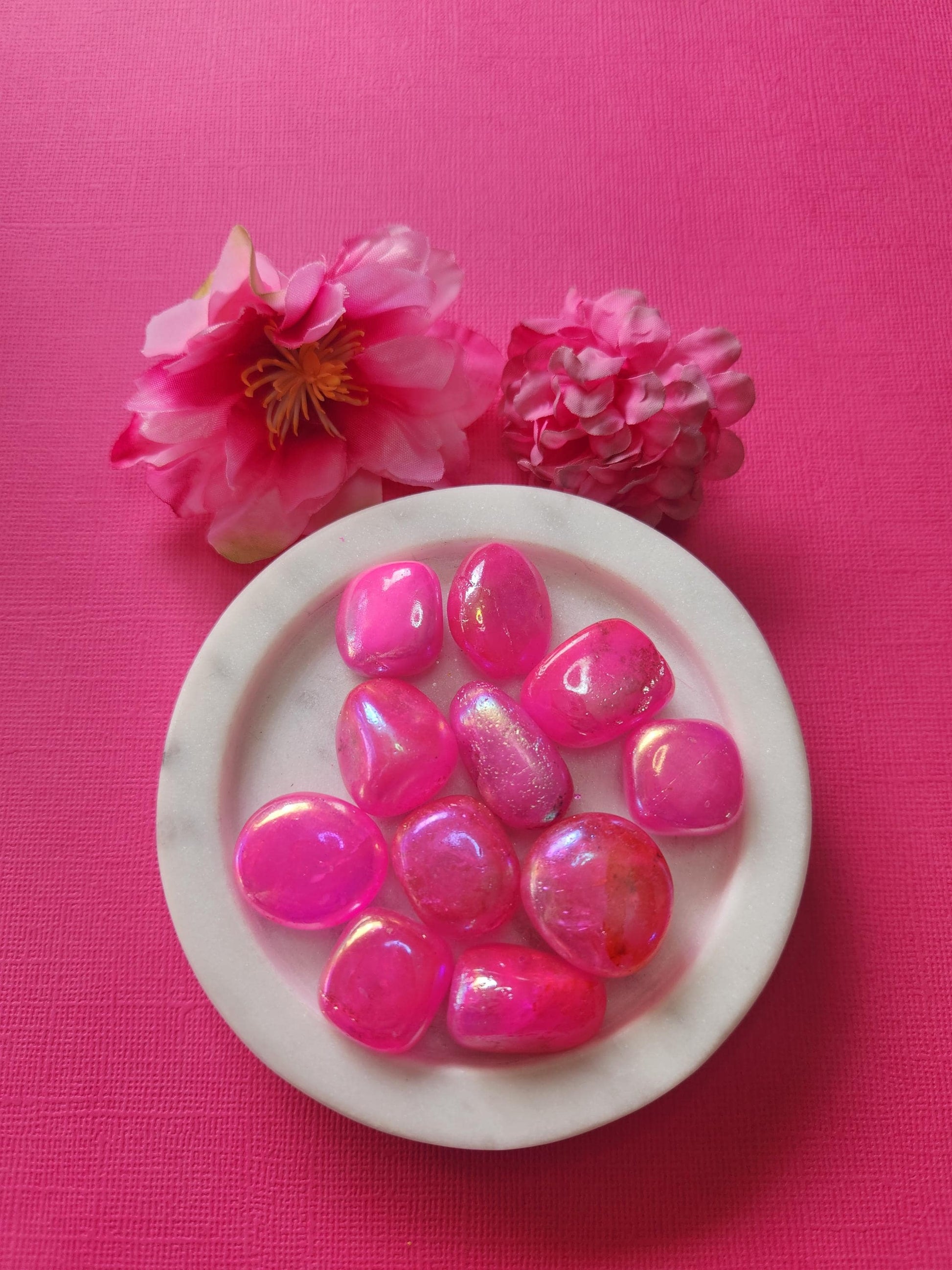 Pink Aura Quartz tumble stones in a white dish with two flower props on a bright pink background. Iridescent pink aura crystals with rainbow sheen, polished to a glossy finish. Healing crystals for love, joy, self-confidence, heart chakra, and spiritual energy. High vibration quartz stones for energy healing, crystal photography, and Etsy crystal shop display.