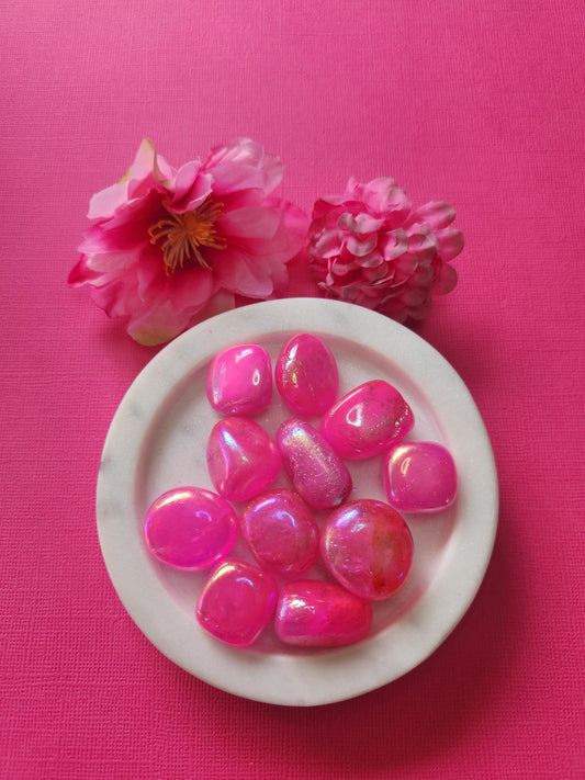Pink Aura Quartz tumble stones in a white dish with two flower props on a bright pink background. Iridescent pink aura crystals with rainbow sheen, polished to a glossy finish. Healing crystals for love, joy, self-confidence, heart chakra, and spiritual energy. High vibration quartz stones for energy healing, crystal photography, and Etsy crystal shop display.