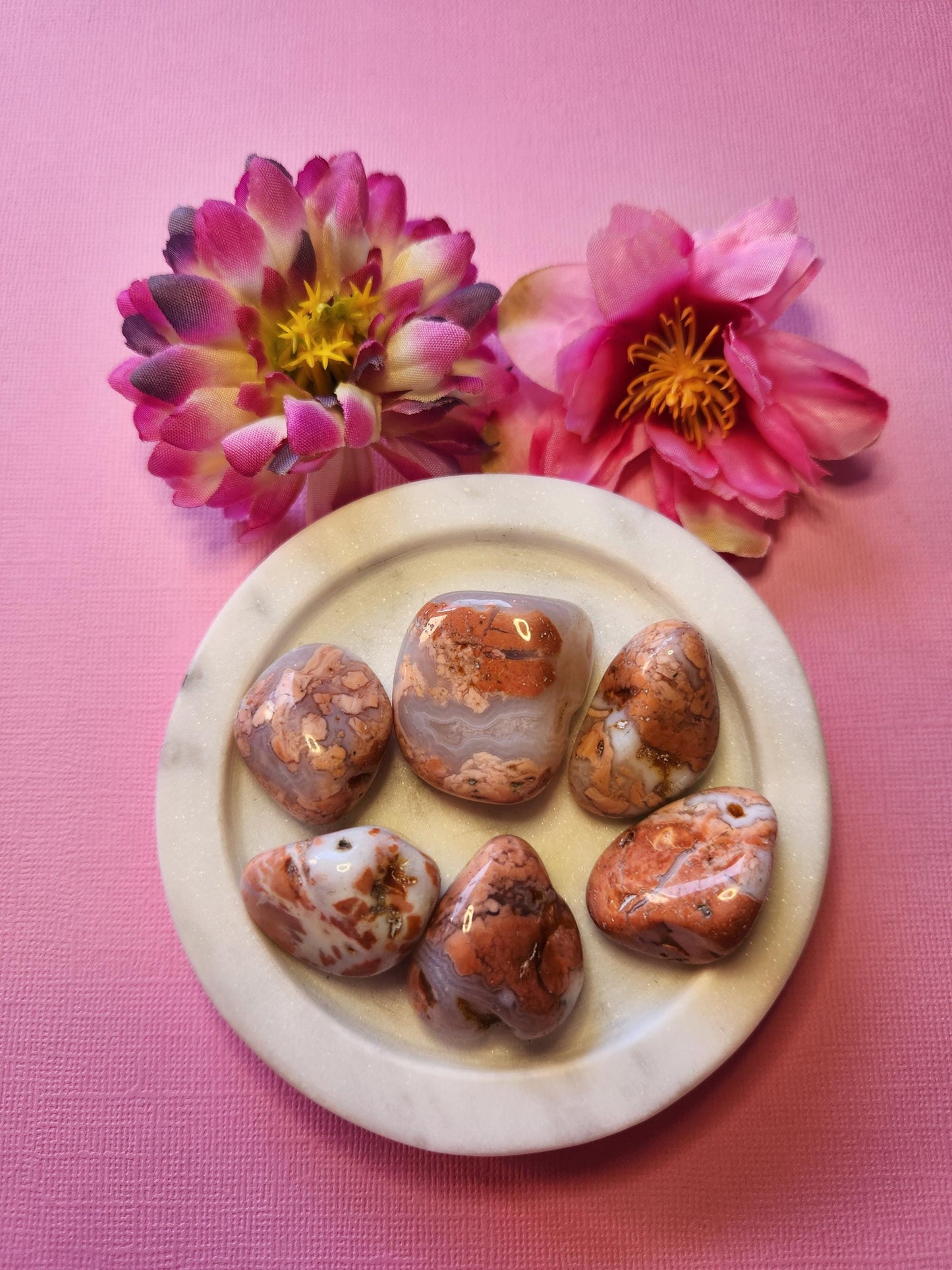 Pink Cotton Candy Agate tumble stones displayed in a white dish with two flower props on a light pink background. Soft pink agate crystals with white banding, polished for a smooth finish. Healing crystals for self love, emotional balance, calm energy, heart chakra, and spiritual wellness. Aesthetic crystal photography for Etsy listing and crystal shop display.