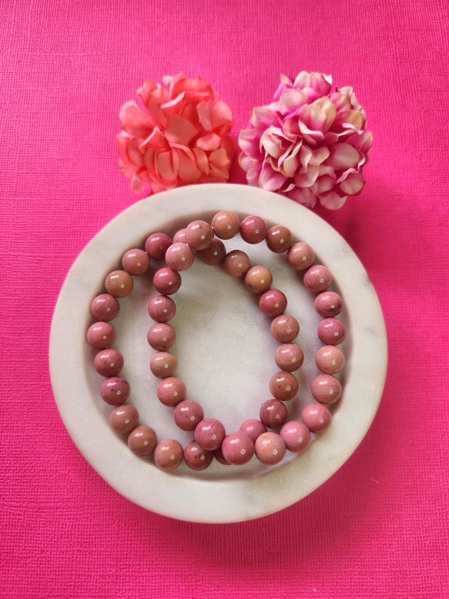 Pink Rhodonite 8mm Beaded Bracelets in white dish with two flower props, pink background, natural gemstone jewelry for emotional healing, love energy, heart chakra support, polished crystal beaded bracelet pair for spiritual wellness and mindful gifting.