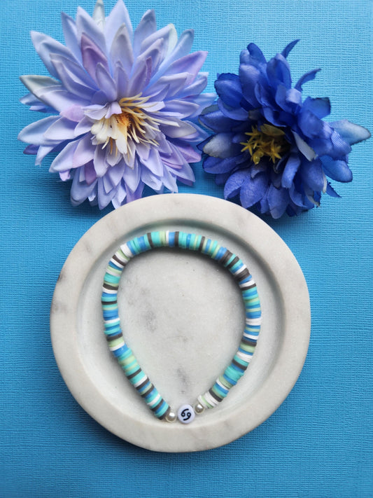 Cancer Zodiac clay beaded bracelet with dark blue, light blue, grey, and white beads, featuring pearl accent beads framing the Cancer charm, handmade astrology jewelry on white dish with flowers, blue background — perfect horoscope gift.