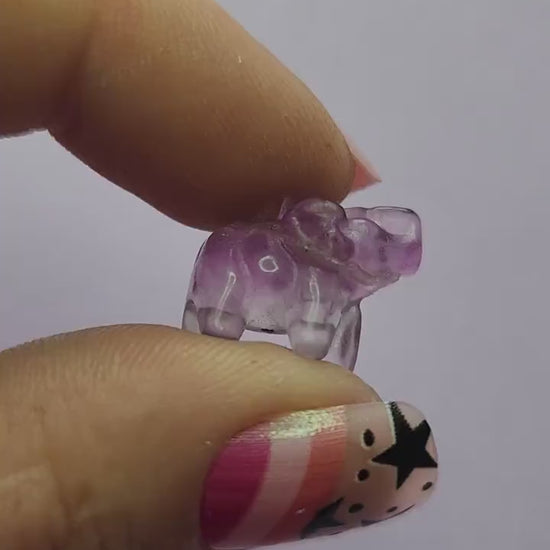 Close-up video of a single fluorite mini elephant carving held in hand, showing polished details and natural purple, green and blue fluorite banding as it catches the light against a lavender backdrop. Perfect for fluorite carvings, mini crystal animals and collectible gemstone elephants