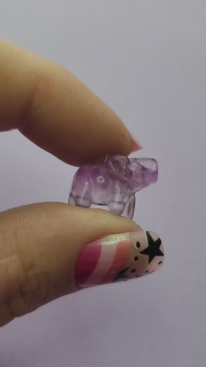 Close-up video of a single fluorite mini elephant carving held in hand, showing polished details and natural purple, green and blue fluorite banding as it catches the light against a lavender backdrop. Perfect for fluorite carvings, mini crystal animals and collectible gemstone elephants