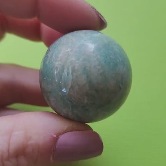 Hand holding a polished Amazonite sphere against a bright green background, highlighting its smooth surface and soothing blue-green colors, great for gifts or home decor