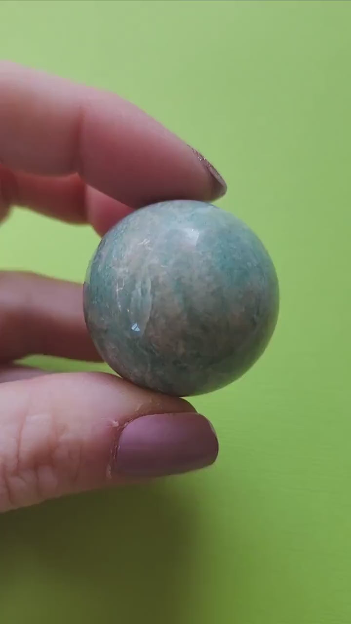 Hand holding a polished Amazonite sphere against a bright green background, highlighting its smooth surface and soothing blue-green colors, great for gifts or home decor