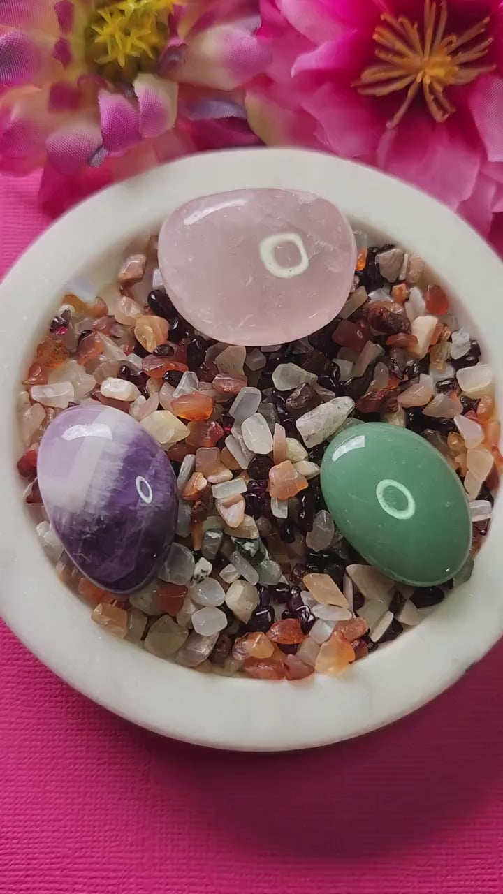 Love Spell Crystal Confetti Mix featuring Carnelian, Peach Moonstone, Garnet, Rose Quartz, Green Aventurine, and Amethyst tumbled stones, displayed on a white dish with two flower props against a soft pink background, ideal for self-love rituals, attraction spells, emotional healing, heart-opening energy, and crystal manifestation practices.