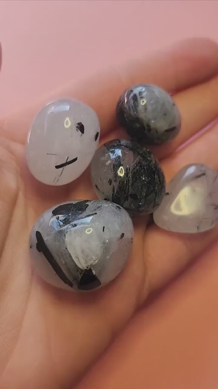 Video of polished Tourmaline in Quartz tumbles held in hand against a pink background, showing clear quartz with black tourmaline inclusions — protective and grounding crystal for clarity, focus, and energy balance.