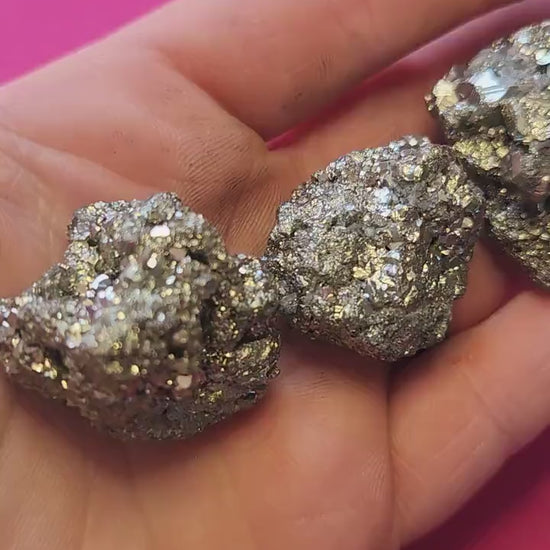 Video of hand holding raw Pyrite crystal pieces against a bright pink background. The metallic gold stones shimmer in the light. Perfect for abundance, manifestation, and crystal collecting