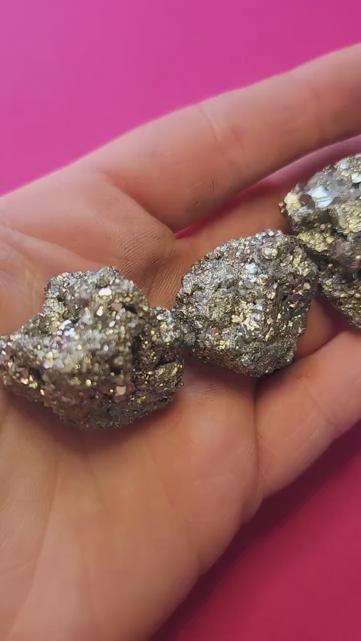Video of hand holding raw Pyrite crystal pieces against a bright pink background. The metallic gold stones shimmer in the light. Perfect for abundance, manifestation, and crystal collecting