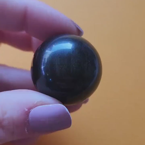Close-up of Gold Sheen Obsidian sphere held in hand, showing its glossy black surface with shimmering golden sheen. Polished crystal sphere for protection, shadow work, meditation, and root chakra energy. Perfect for spiritual decor, energy work, and crystal collectors.