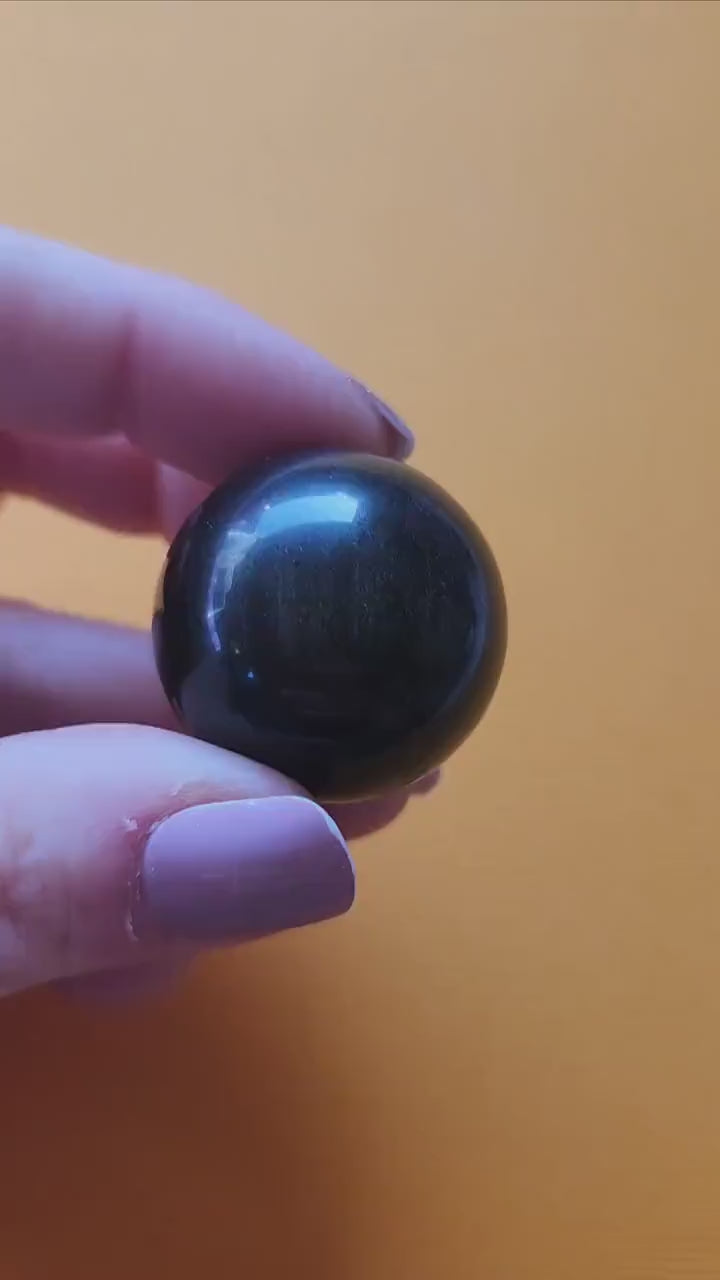 Close-up of Gold Sheen Obsidian sphere held in hand, showing its glossy black surface with shimmering golden sheen. Polished crystal sphere for protection, shadow work, meditation, and root chakra energy. Perfect for spiritual decor, energy work, and crystal collectors.