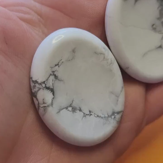 Polished Howlite worry stone held in hand, showing soft white color with grey veining, thumb groove visible, on a bright yellow background. Stress relief, meditation, calming crystal.