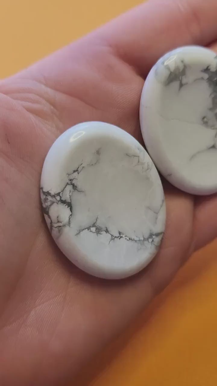 Polished Howlite worry stone held in hand, showing soft white color with grey veining, thumb groove visible, on a bright yellow background. Stress relief, meditation, calming crystal.