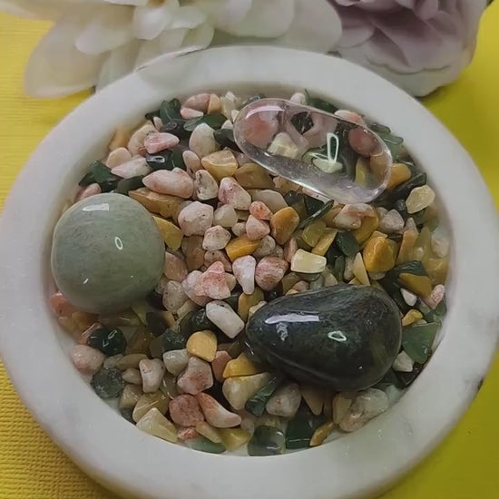 Abundance Spell Crystal Confetti Mix featuring Yellow Aventurine, Sunstone, Green Aventurine, Moss Agate, Clear Quartz, and Amazonite tumbled stones, 50g crystal mix displayed on a white dish with two flower props against a vibrant yellow background, ideal for abundance rituals, wealth manifestation grids, prosperity crystal magic, and spiritual decor.