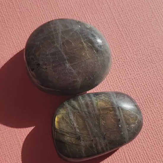 Close-up of two polished Labradorite palm stones held to showcase pink and yellow flashes against a light pink backdrop. Natural Labradorite crystals ideal for meditation, aura protection, chakra balancing, Reiki, spiritual practice, pocket carrying, and crystal collections. Perfect for crystal enthusiasts, energy work, and thoughtful gifts.