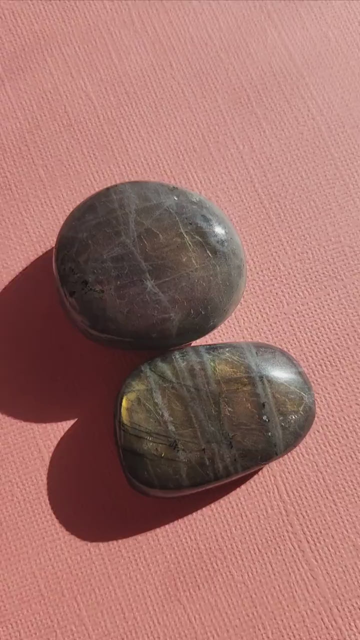 Close-up of two polished Labradorite palm stones held to showcase pink and yellow flashes against a light pink backdrop. Natural Labradorite crystals ideal for meditation, aura protection, chakra balancing, Reiki, spiritual practice, pocket carrying, and crystal collections. Perfect for crystal enthusiasts, energy work, and thoughtful gifts.