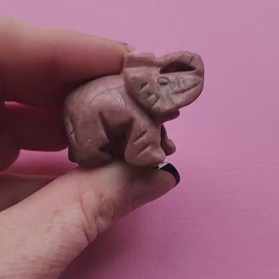 Video of a Rhodonite crystal carving held in hand against a light pink background, showing smooth pink and black stone details — loving crystal decor for emotional healing, self-love, and heart chakra energy