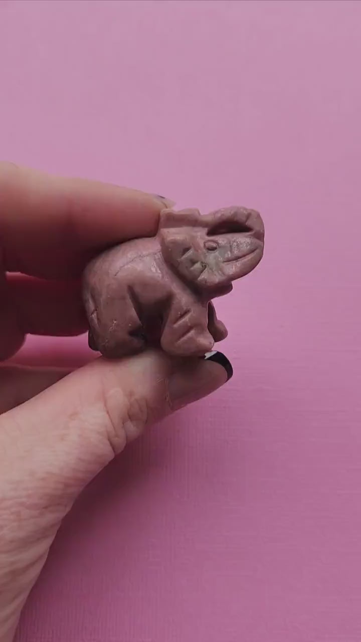 Video of a Rhodonite crystal carving held in hand against a light pink background, showing smooth pink and black stone details — loving crystal decor for emotional healing, self-love, and heart chakra energy