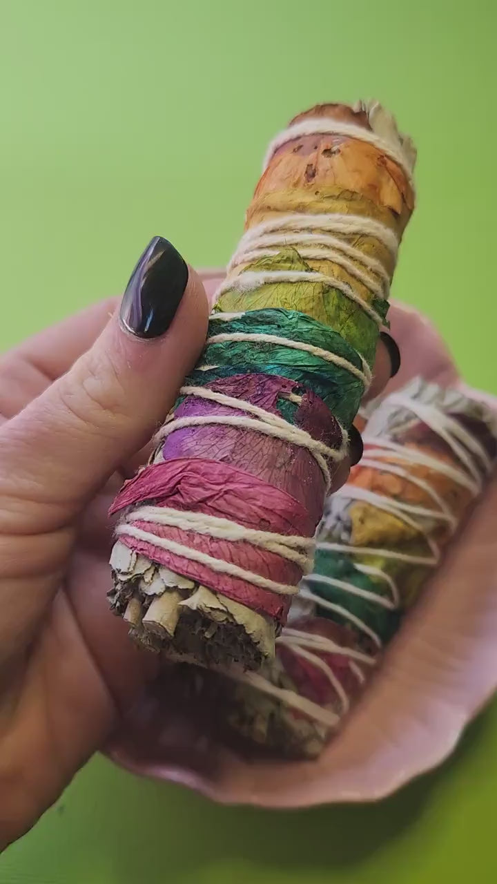 Video of a White Sage smudge stick with chakra petals held in hand against a green background, with more sticks displayed in a pink flower bowl behind — cleansing and purifying ritual sage for energy clearing, chakra balancing, and spiritual wellness.