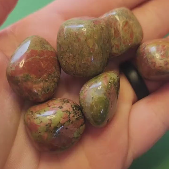 Video of polished Unakite tumbles held in hand against a green background, showing green and pink stone patterns — nurturing and balancing crystal for emotional healing, harmony, and grounding.