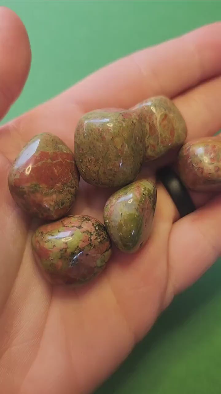 Video of polished Unakite tumbles held in hand against a green background, showing green and pink stone patterns — nurturing and balancing crystal for emotional healing, harmony, and grounding.