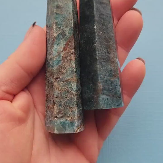 Hands holding and showcasing two polished Blue Apatite towers against a light blue background, highlighting their smooth finish and vibrant blue color.