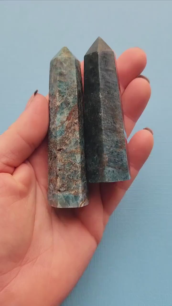 Hands holding and showcasing two polished Blue Apatite towers against a light blue background, highlighting their smooth finish and vibrant blue color.